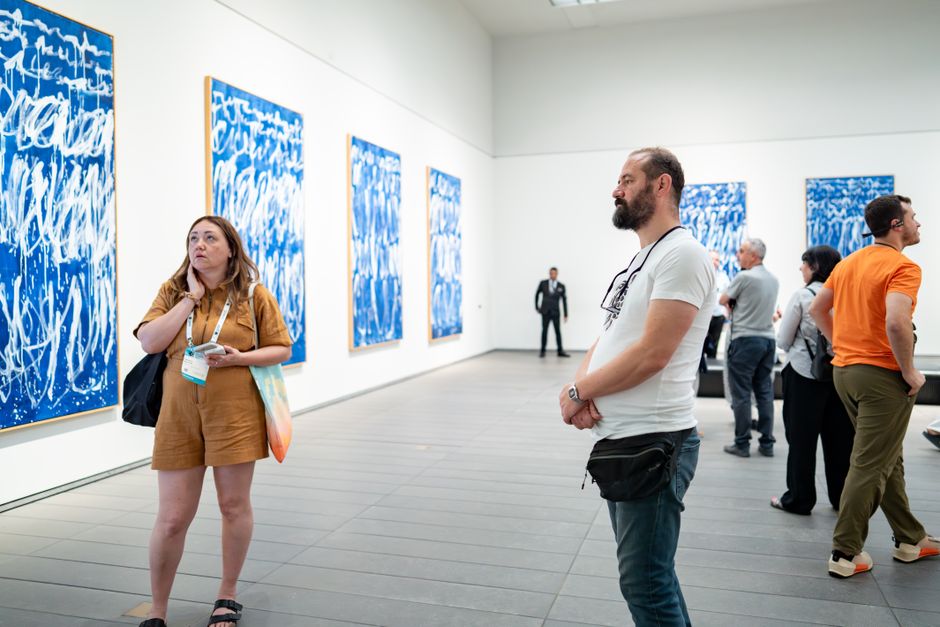 アートギャラリーで青い抽象画に魅了される来場者たち。現代アートが展示された屋内で撮影。多様な来場者がその光景を彩っています。