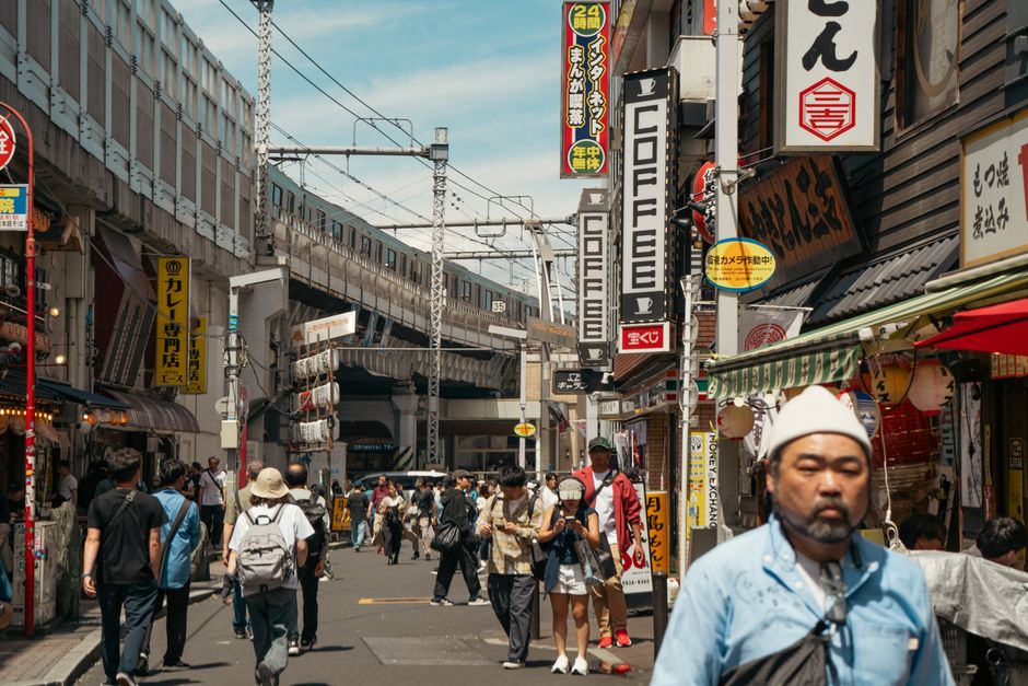 活気あふれる東京の街並み。人々、商店、そして頭上を走る電車が描かれている。