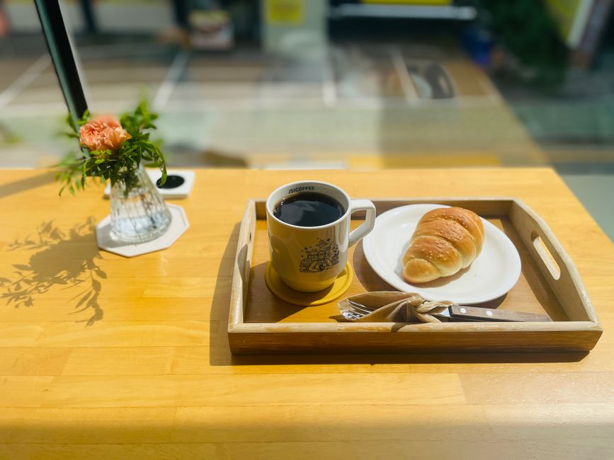 日当たりの良い木製のテーブルにコーヒー、クロワッサン、花瓶が置かれた、温かみのある居心地の良いカフェの雰囲気。