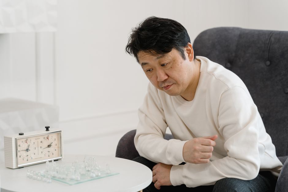室内でチェスのゲームを考えるアジア人男性、白いセーターを着ており、近くにアナログ時計があります。